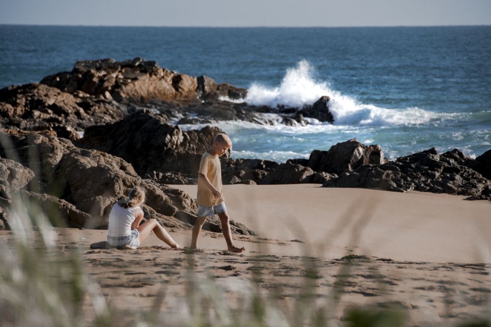 2 people walking along beach with waves and rocks behind them.